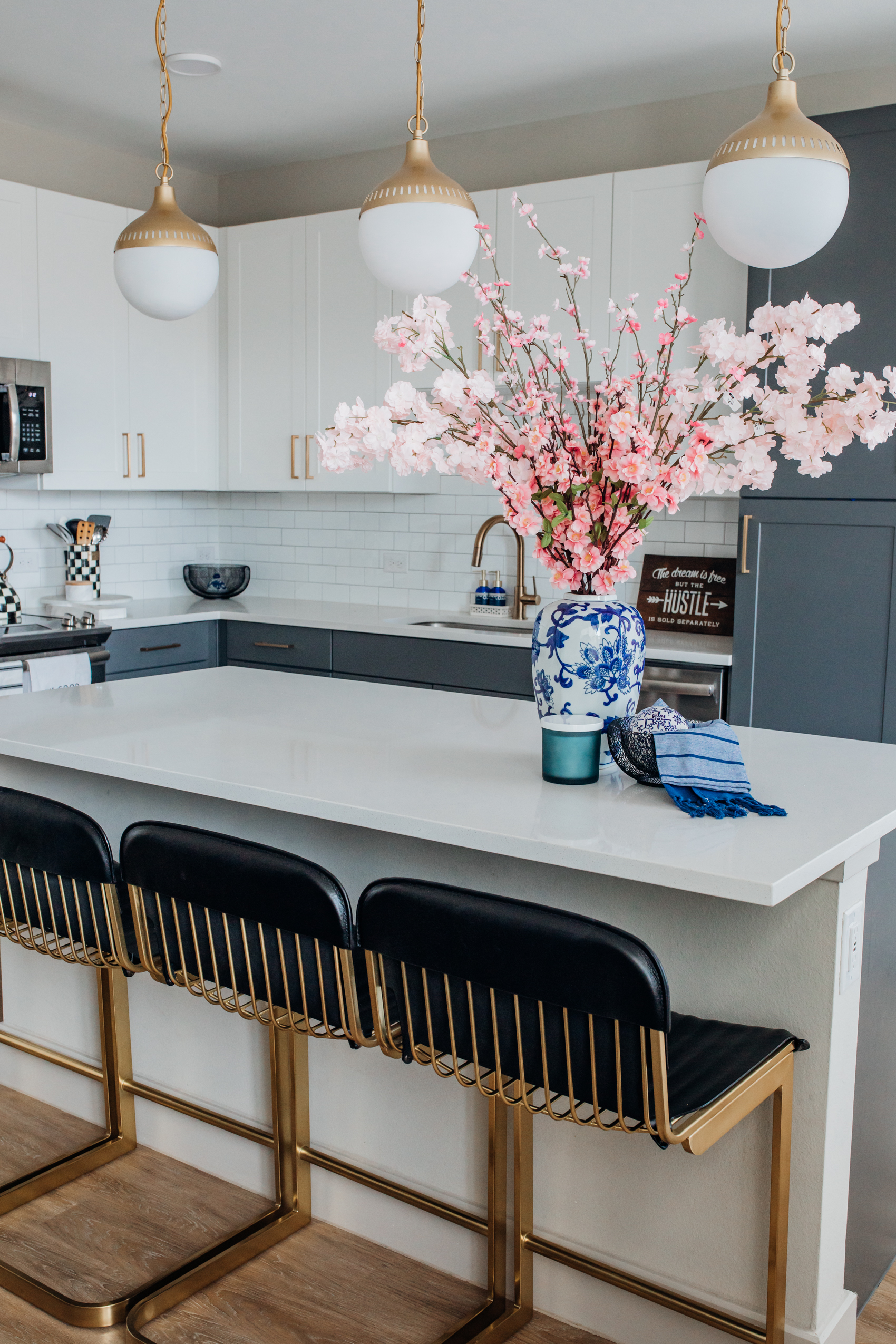 Spring home decor in a white and blue two-toned kitchen with CB2 Rake black leather barstools, gold pendants, subway tile and cherry blossom arrangements and black mesh bowls