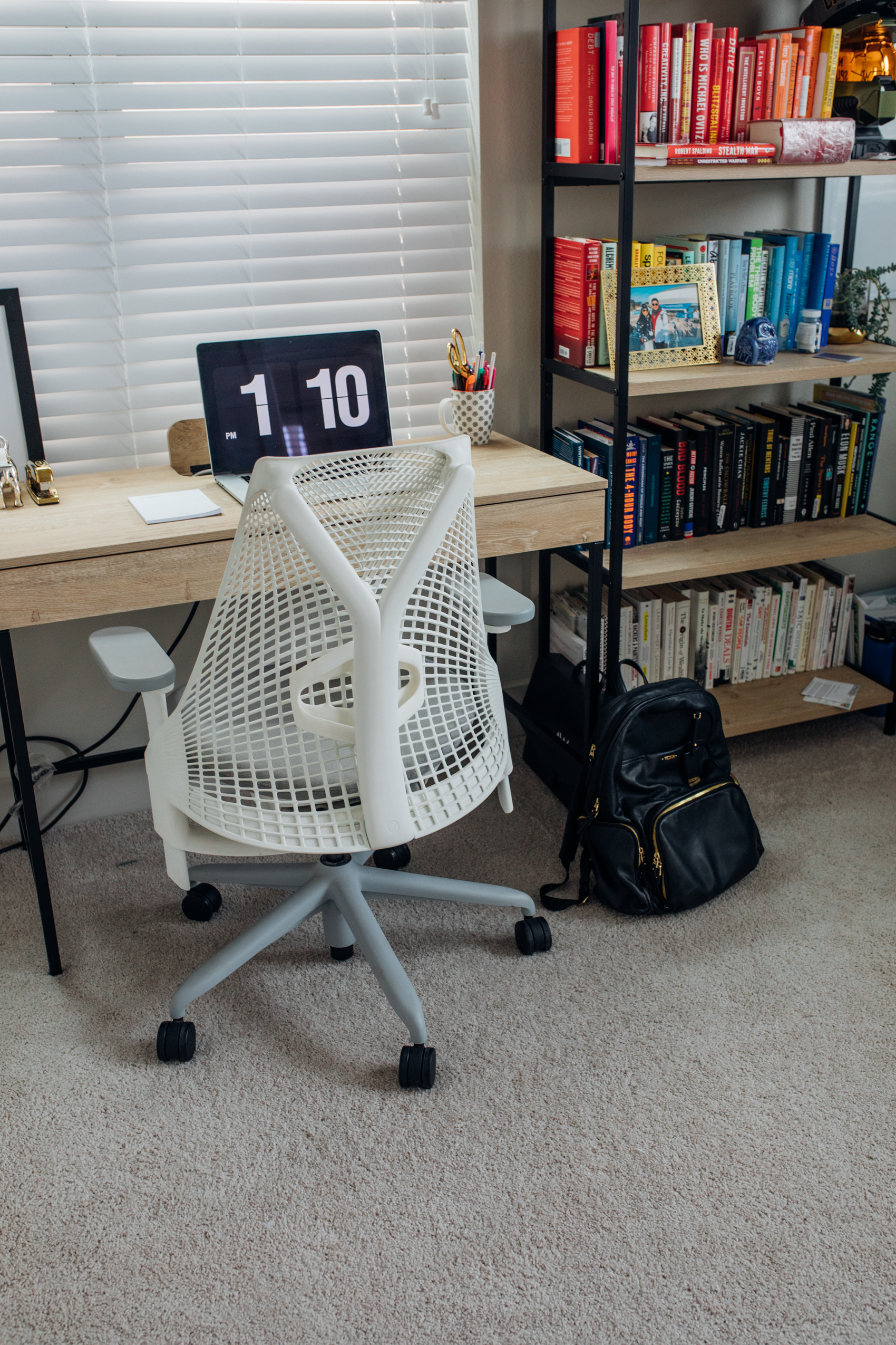 Dallas home office with writing desk, Herman Miller white Sayl chair, Tumi leather backpack and colorful bookshelf