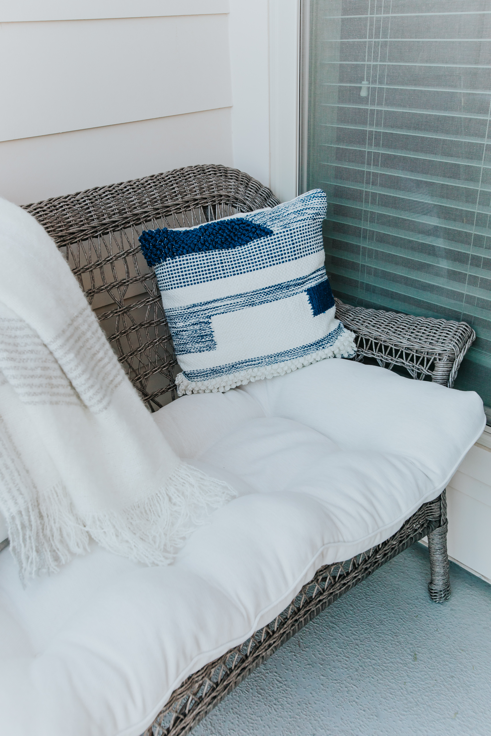 Apartment patio space with a gray wicker settee, white herringbone cushion, blue throw pillow and gray striped blanket