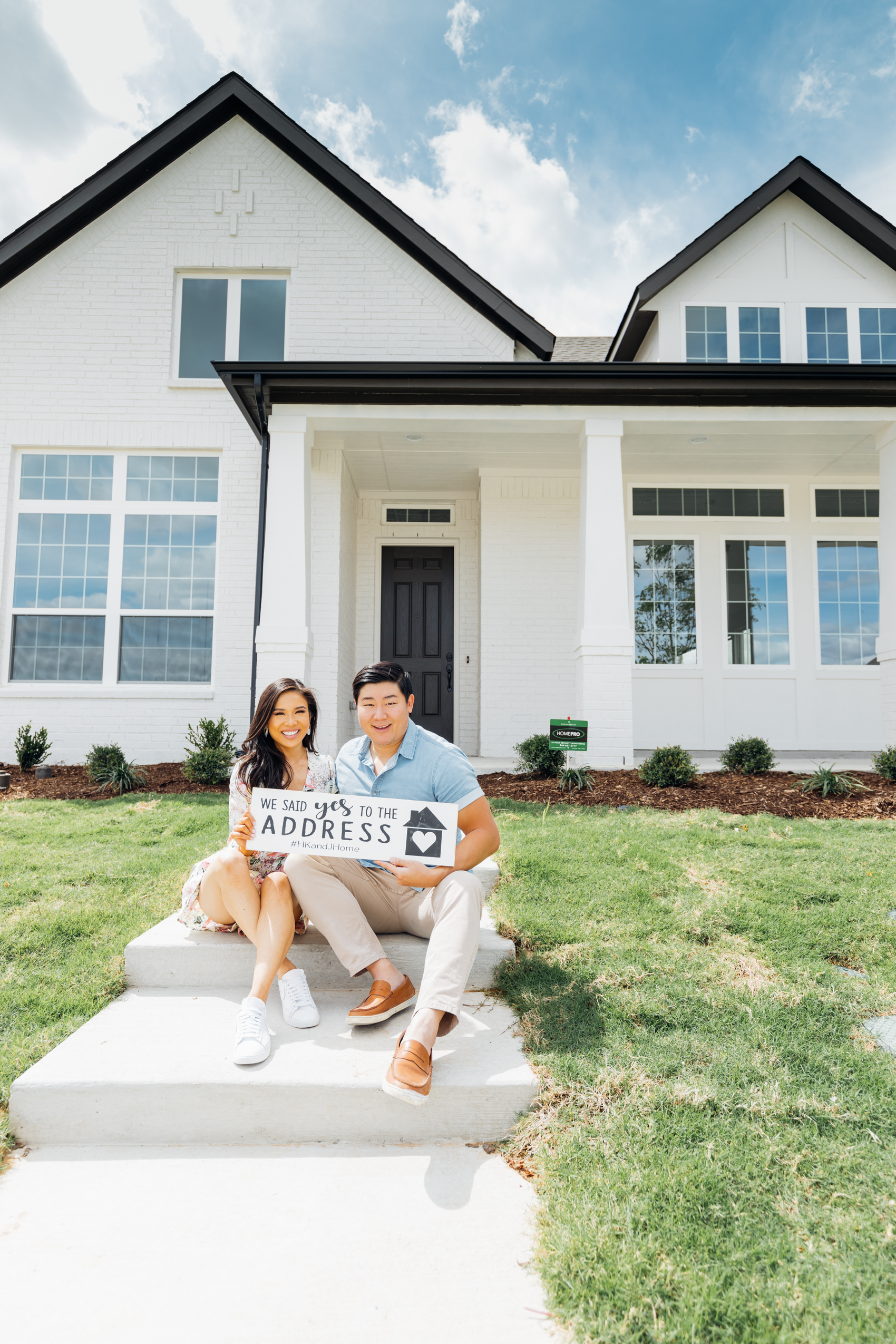 Blogger Hoang-Kim and her fiance Johnny in front of their new build home in Dallas that's a one-story home with white painted brick and black trim wearing a floral romper and chambray shirt
