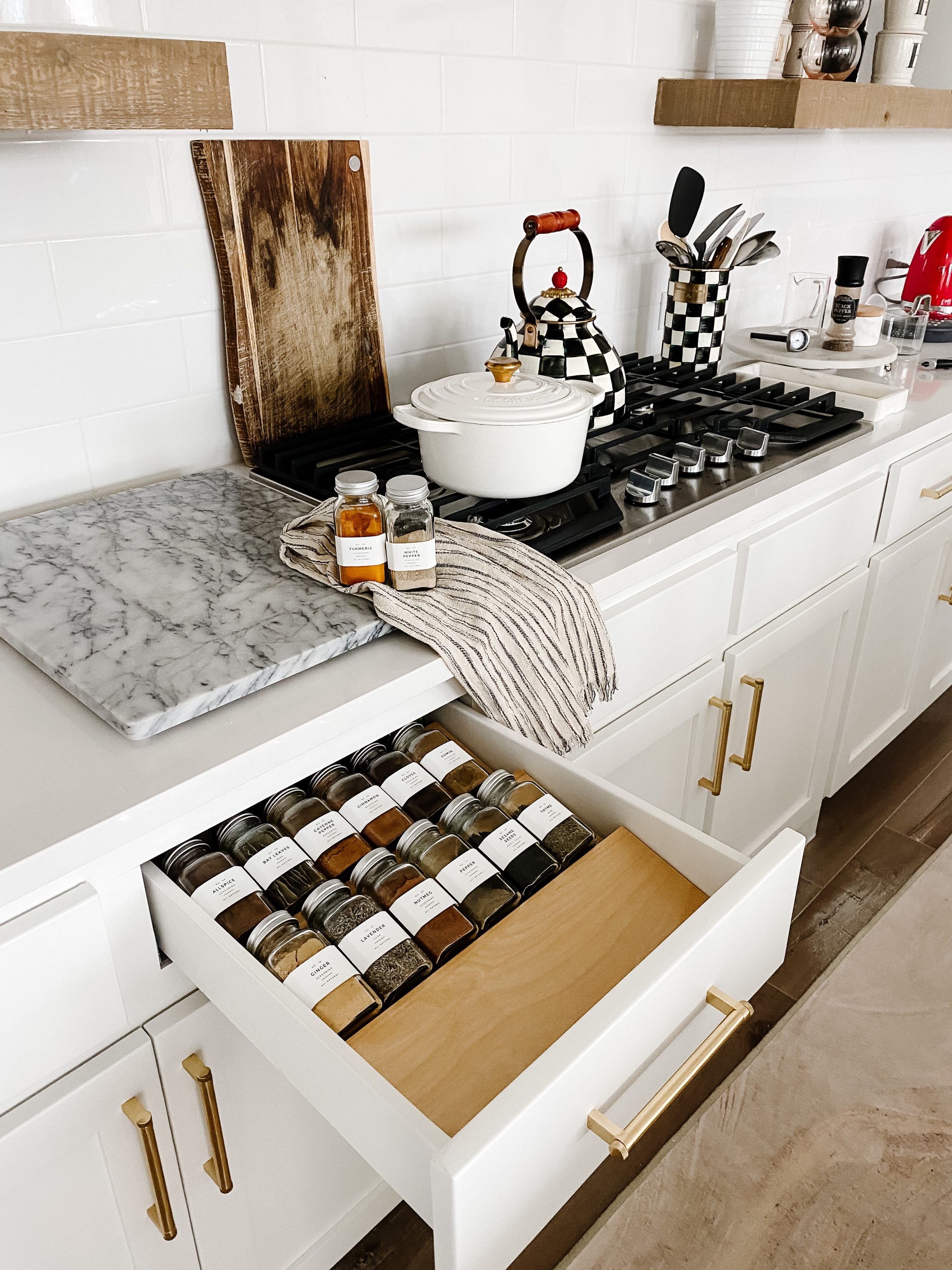 Spice drawer organization with modern labels in a white kitchen with shaker cabinets, white Le Creuset dutch oven with gold knob and MacKenzie-Childs kettle and utensil holder