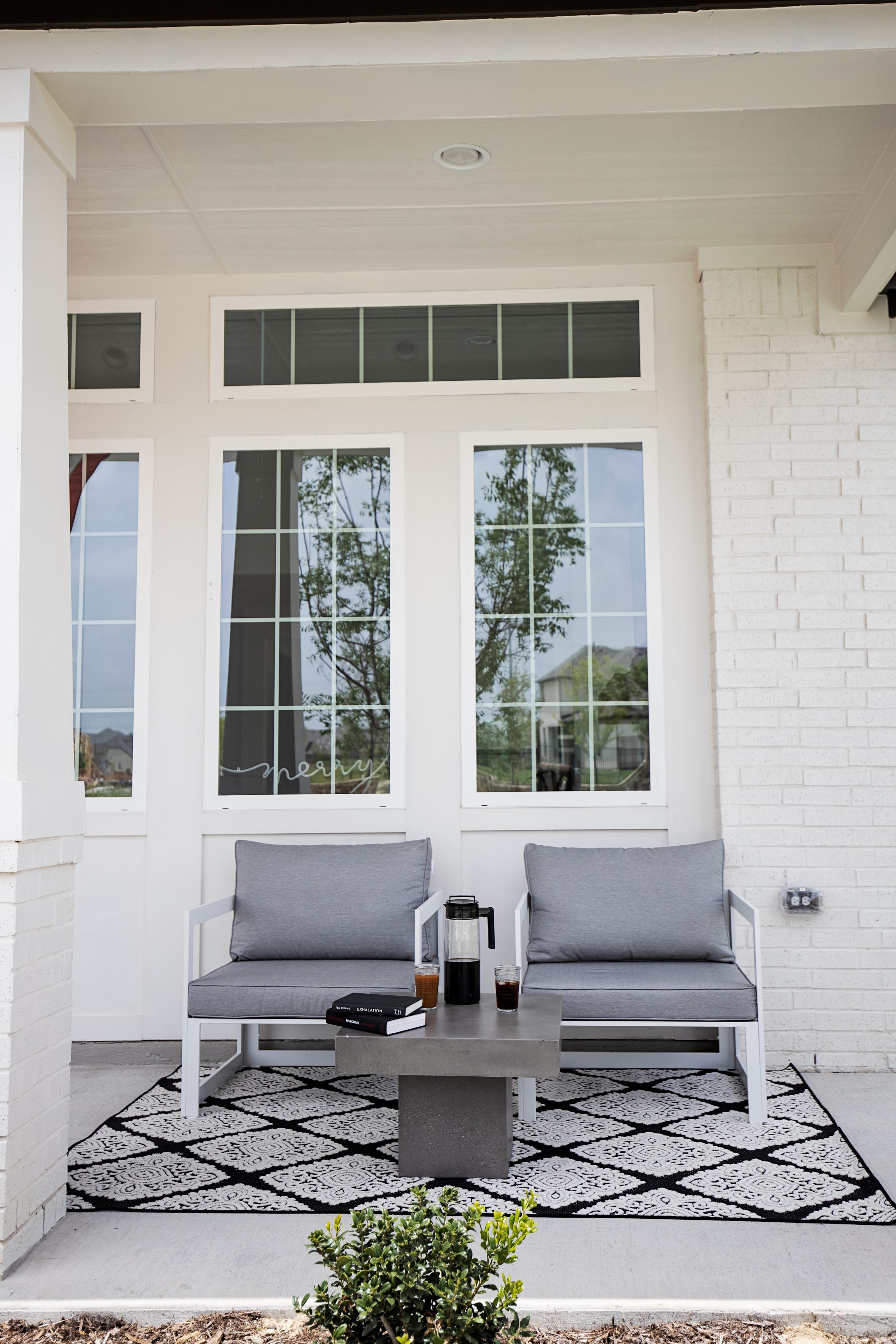 Modern outdoor furniture with white and gray chairs, concrete table, black and white rug on a white painted brick home front patio