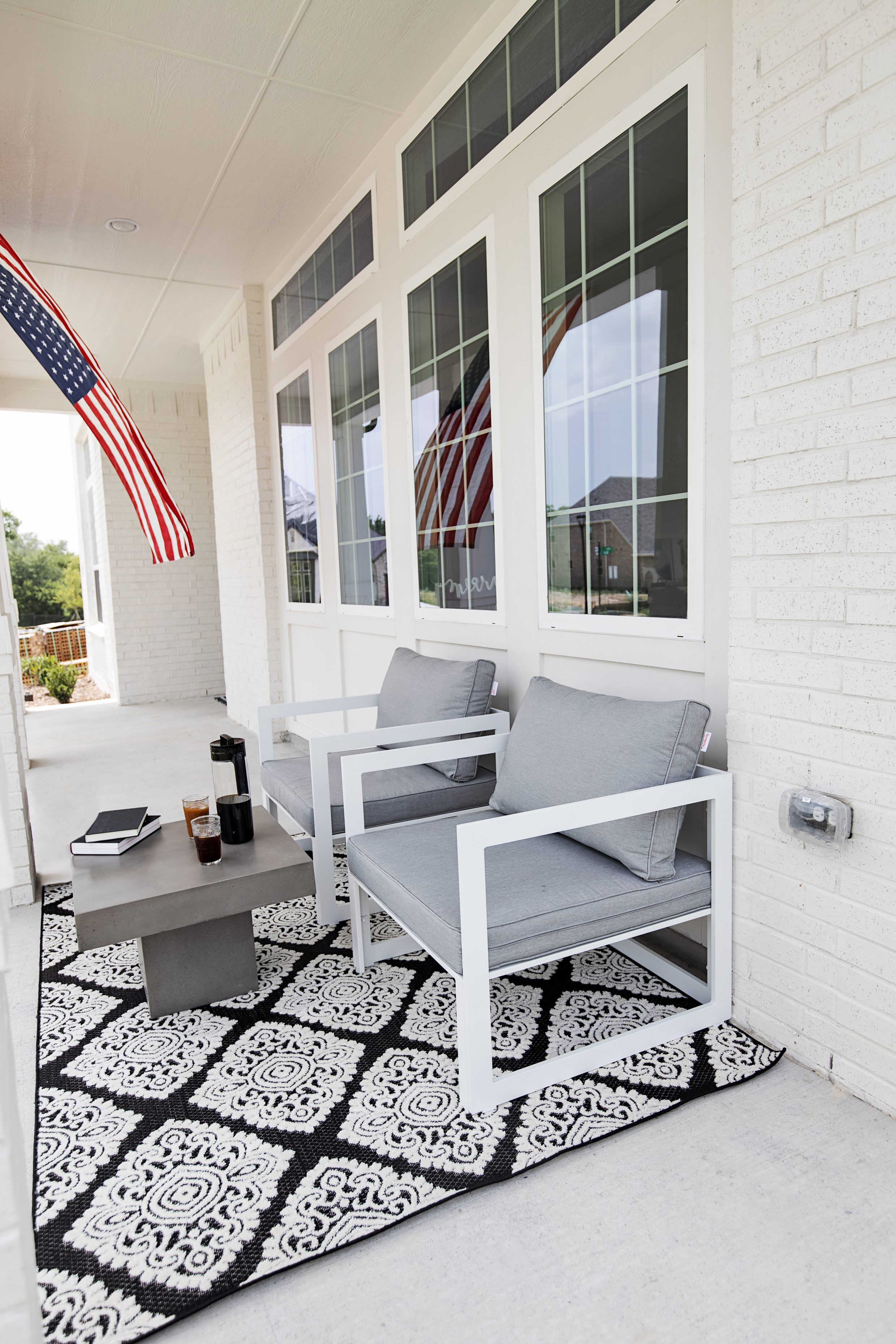 Modern outdoor furniture on front yard patio with white and gray chairs, concrete table, black and white rug and American flag on a white painted brick home