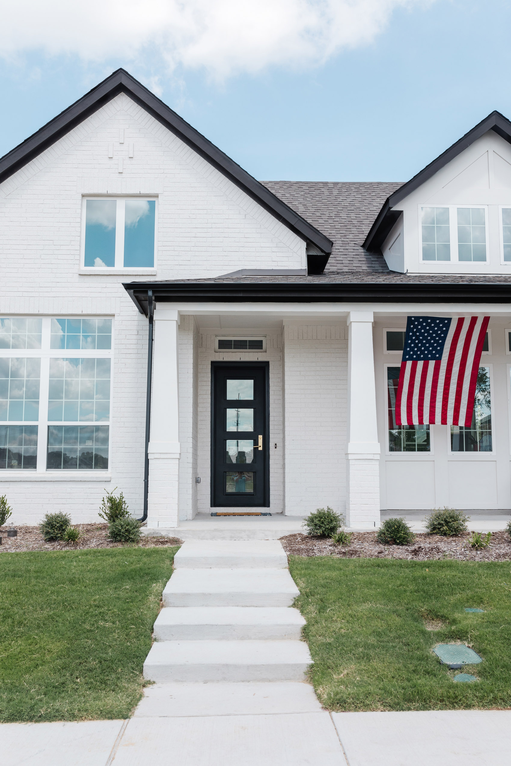 Transitional home with an iron front door, white painted brick, black trim and American flag on the front porch