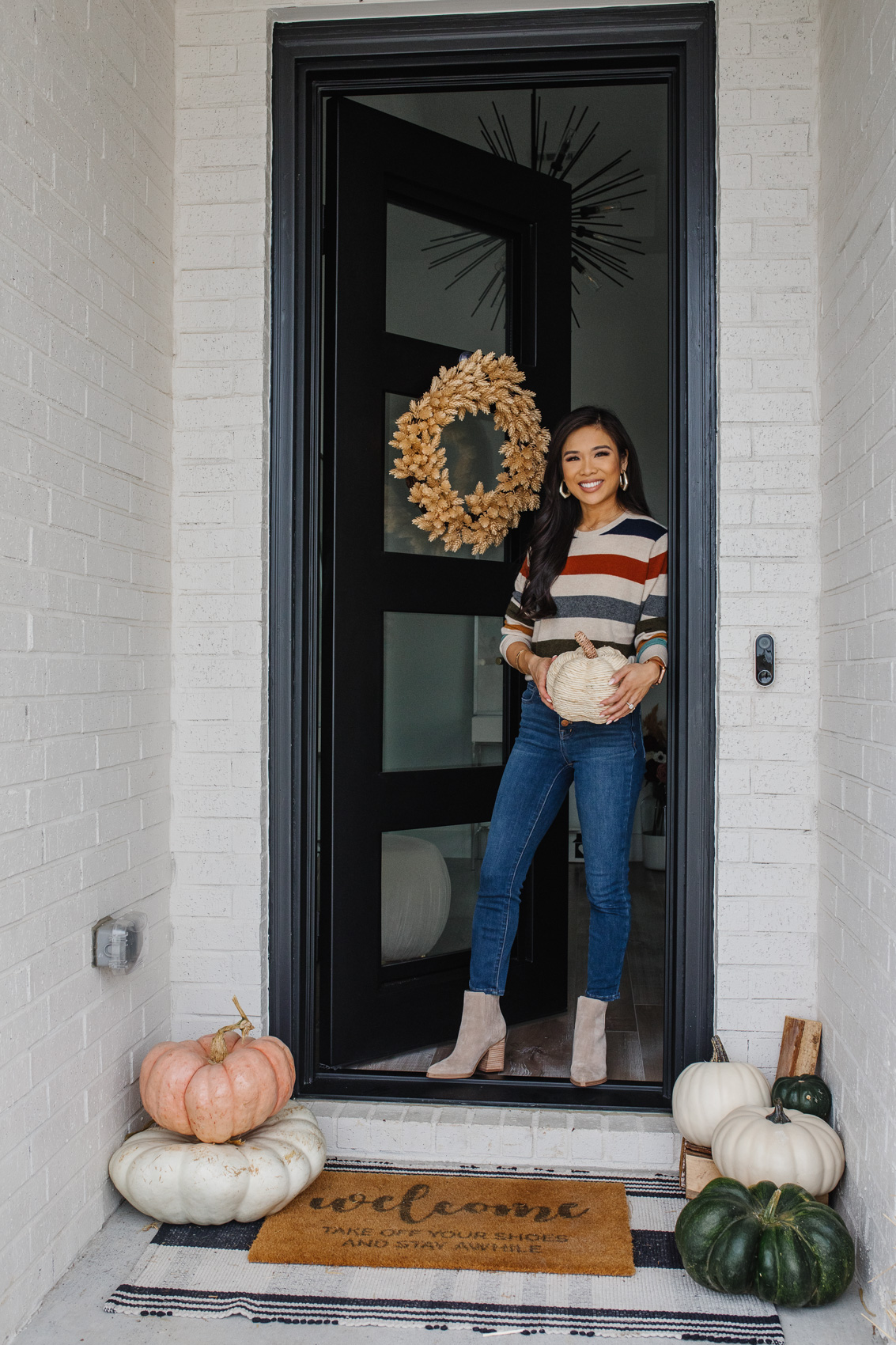 Blogger Hoang-Kim on her fall front porch with an iron door, neutral pumpkins, plaid rug, doormat, hops wreath wearing a cashmere striped sweater, blue jeans and gold earrings