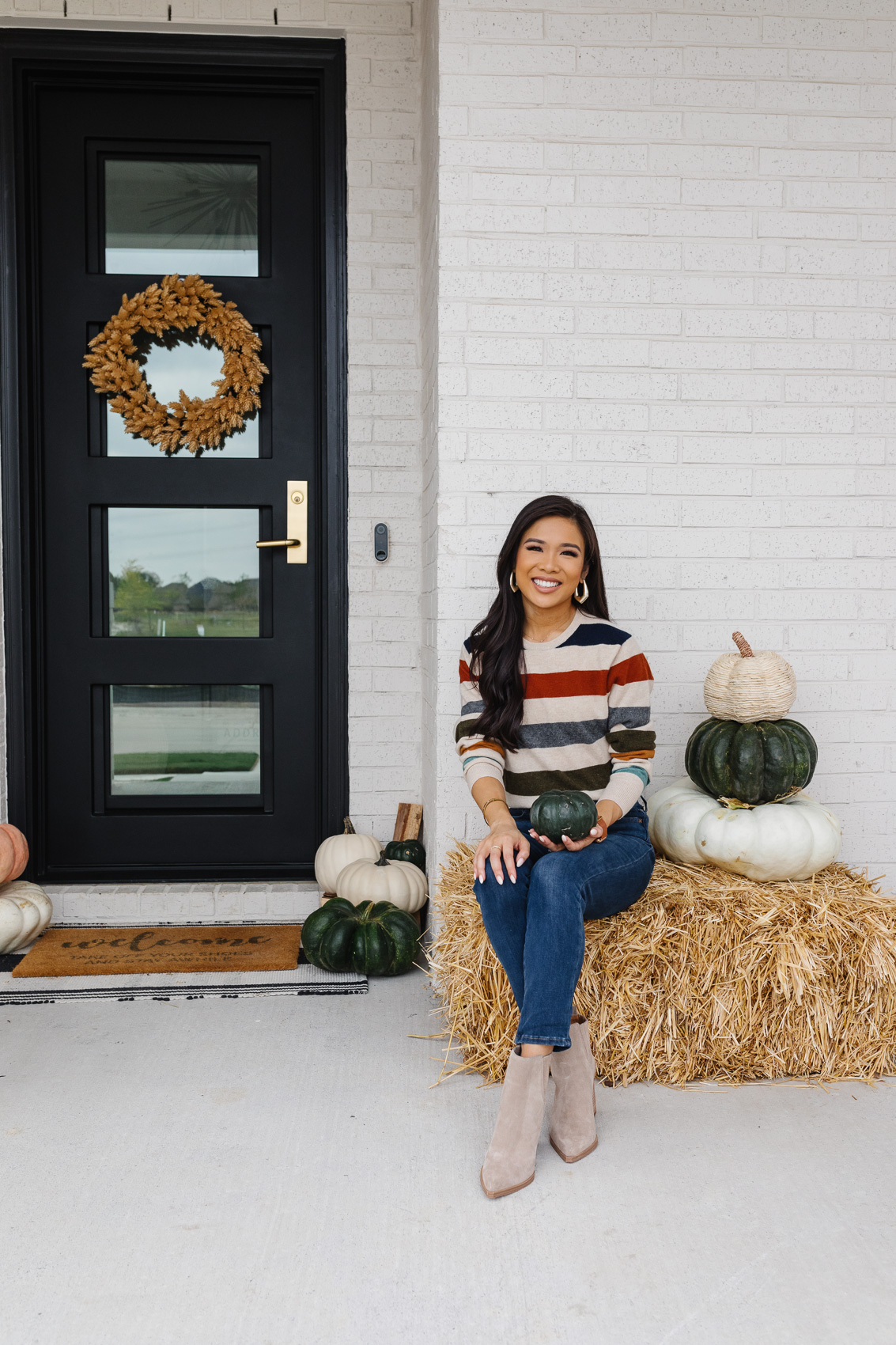 Blogger Hoang-Kim wearing a striped cashmere sweater and blue jeans on her fall front porch with white, green and pastel pumpkins and iron front door