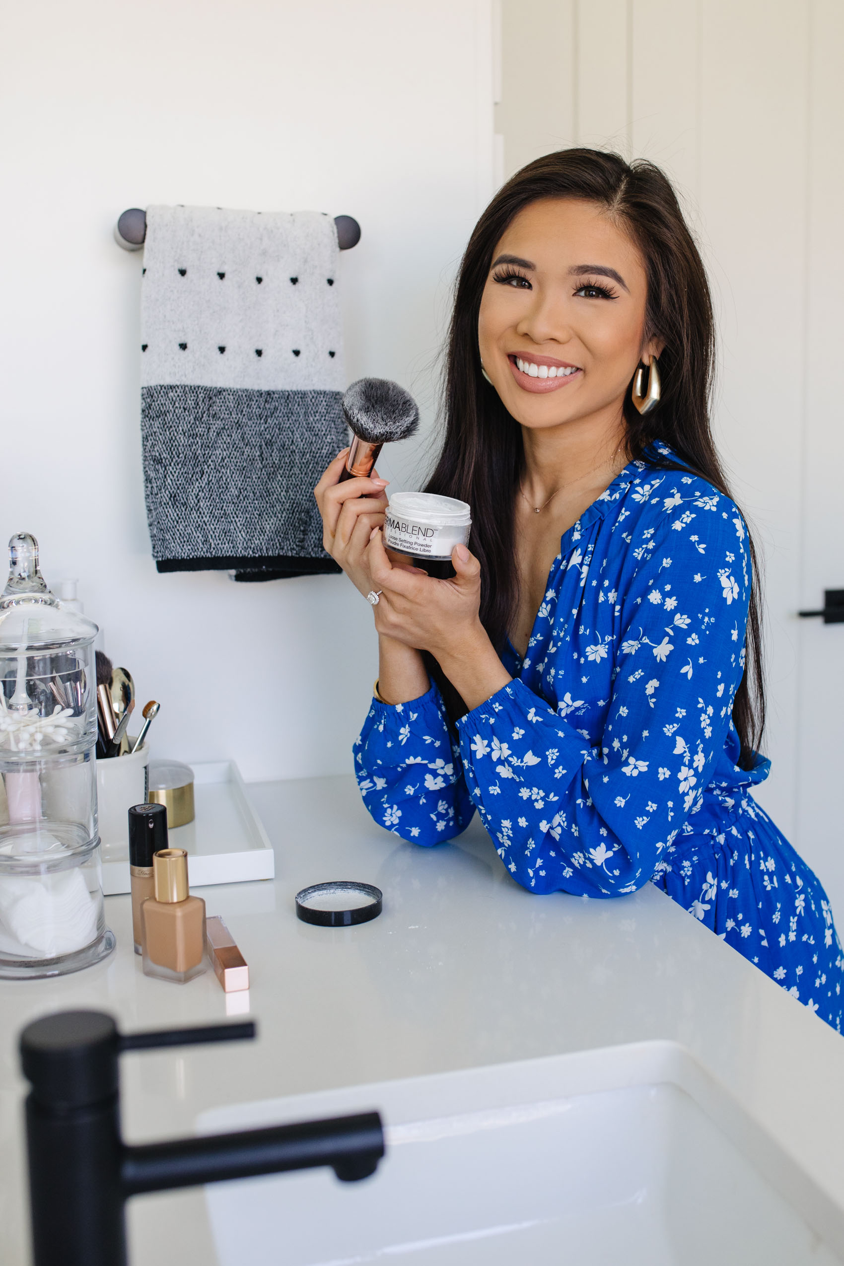 Beauty blogger Hoang-Kim applying Dermablend loose setting powder in her master bathroom with white marble countertops wearing a blue Draper James dress with gold Kendra Scott Earrings