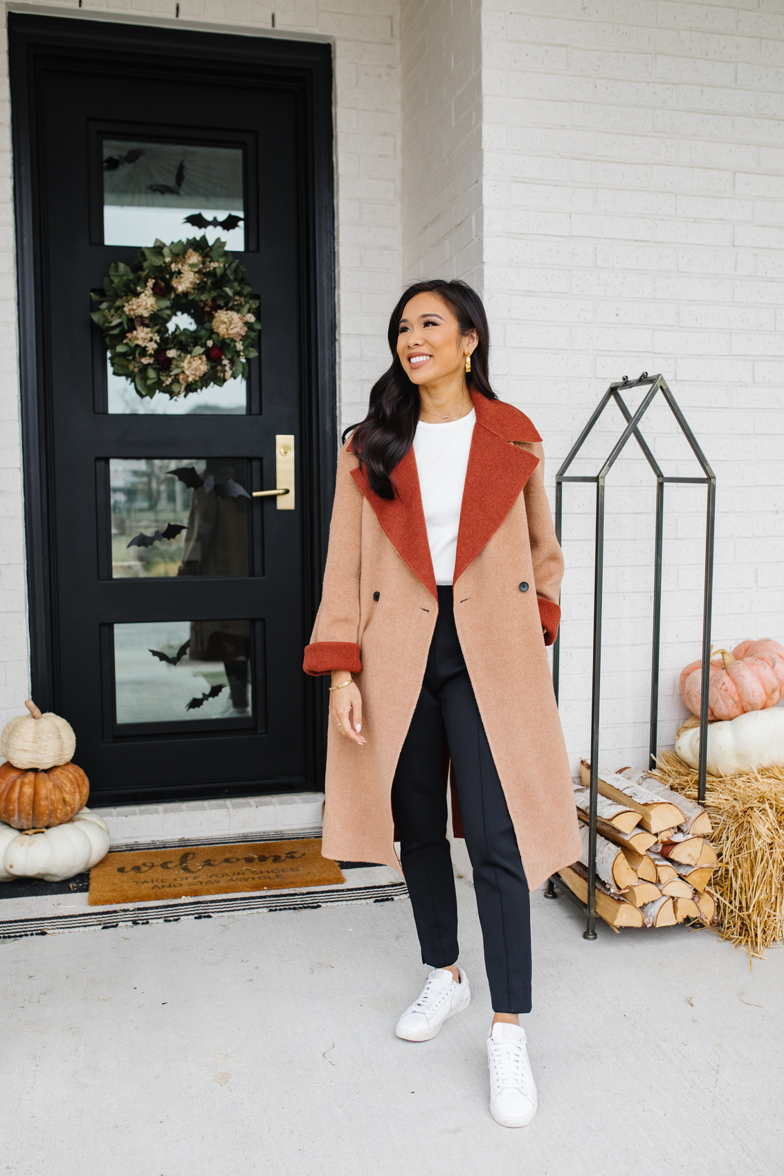 Blogger Hoang-Kim wears a camel coat made of wool by M.M.LaFleur with black pants, white sneakers and a white top in front of her fall front porch