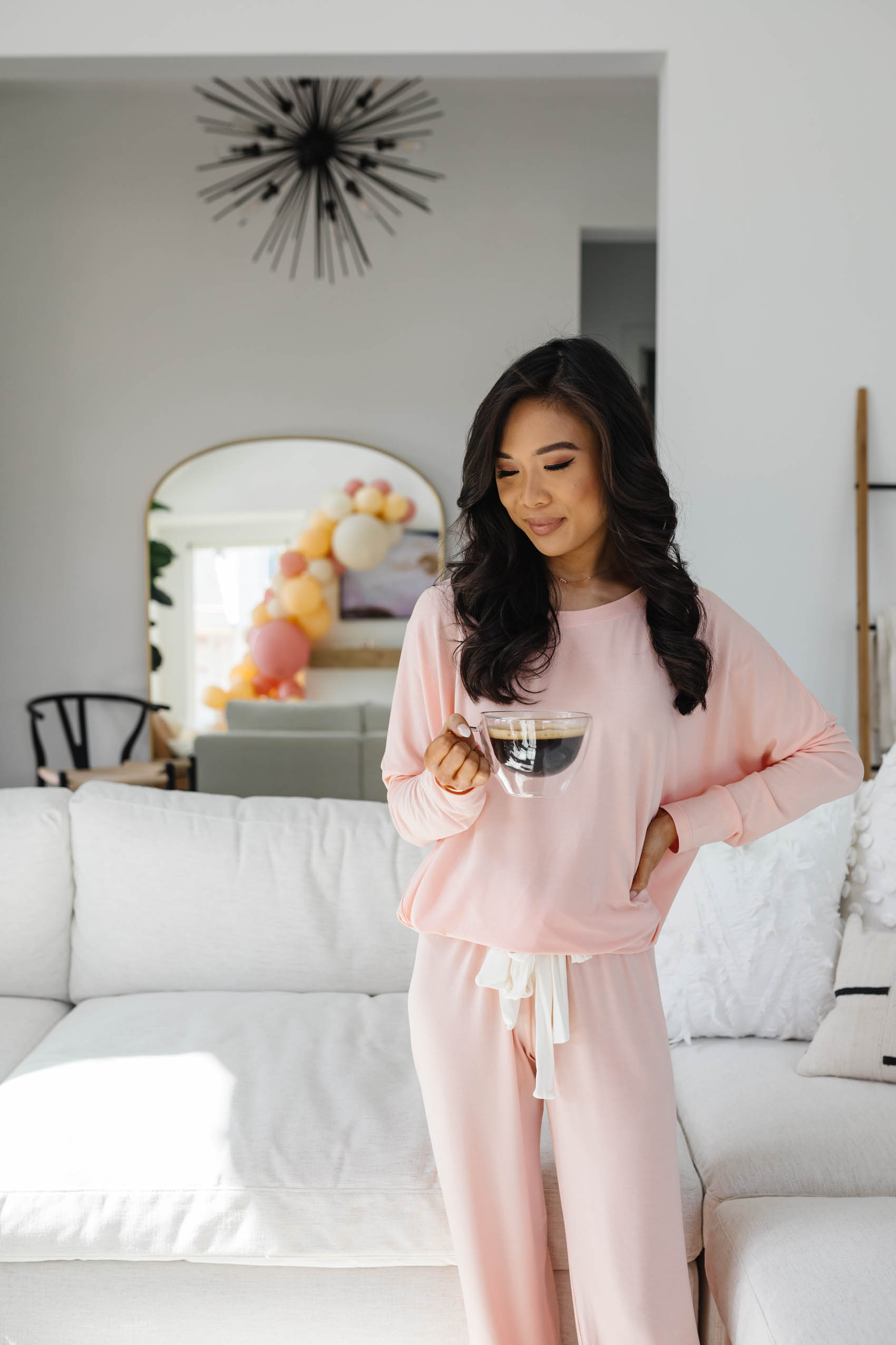 Blogger Hoang-Kim wears a pink eberjey pajama set holding a glass coffee mug in her living room decorated with an Arhaus Beale sofa, rejuvenation arch mirror in her Dallas home