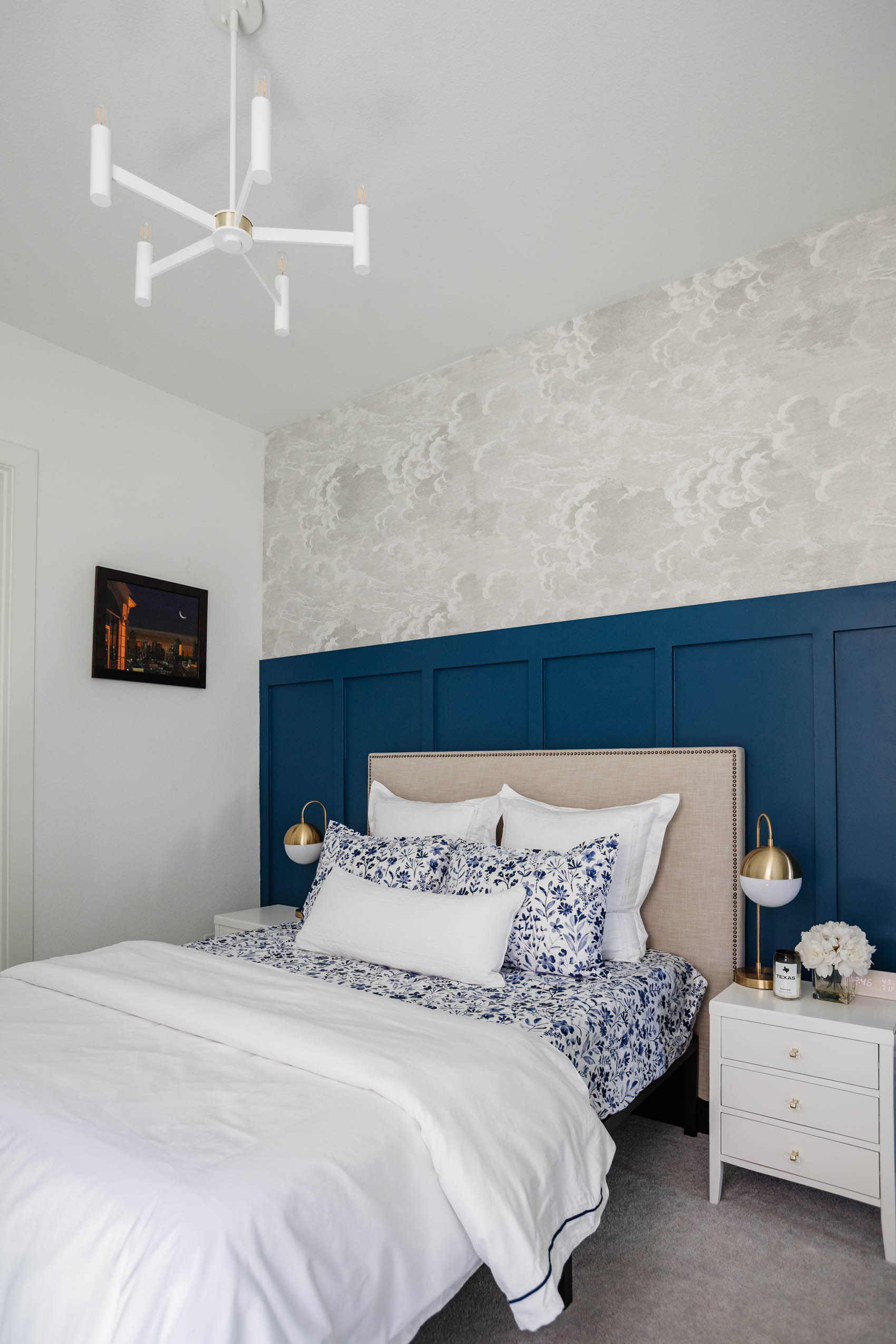 Guest bedroom with board and batten in PPG Chinese Porcelain with Cole & Son nuvolette wallpaper, west elm upholstered headboard, boll & branch botanical print bedsheets, white nightstands and chandelier and brass table lamps