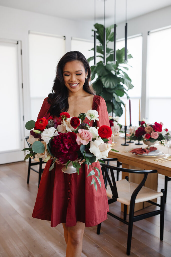 Blogger Hoang-Kim holding a fall floral arrangement with red peonies, ranunculus, toffee roses, roses wearing a rust colored Anthropologie dress in front of her McGee & Co Antoni extendable dining table set for Thanksgiving Galsgiving