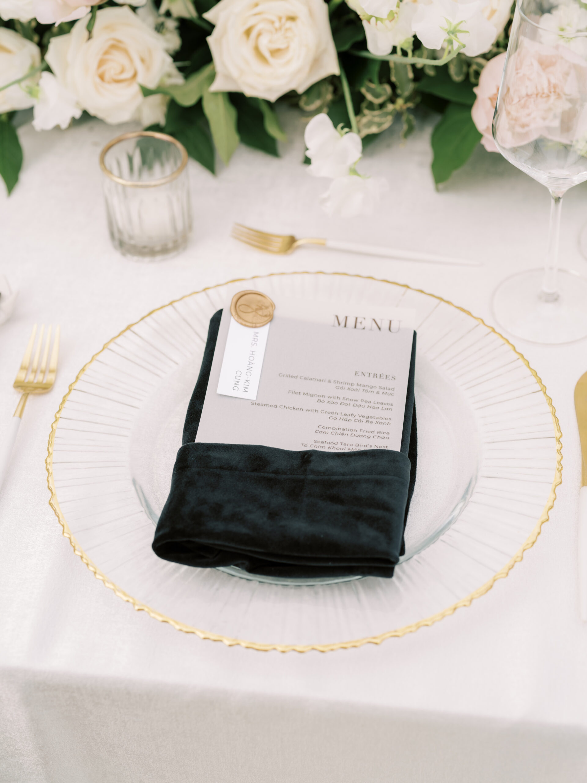 Modern and timeless wedding place setting with pleated gold rimmed charger, black velvet napkin, white and gold cutipol flatware, gold seal place card holder and lush florals