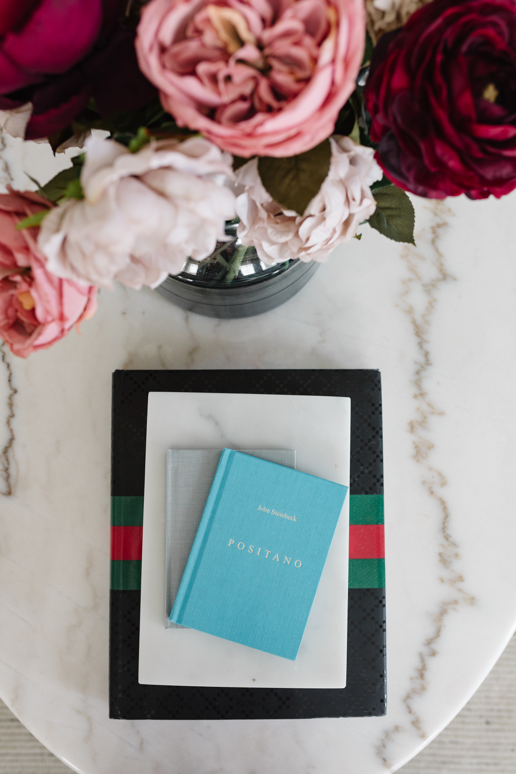 Positano by John Steinbeck linen bound book along with a white marble box to store knick knacks, Gucci book and a faux floral arrangement for coffee table styling.