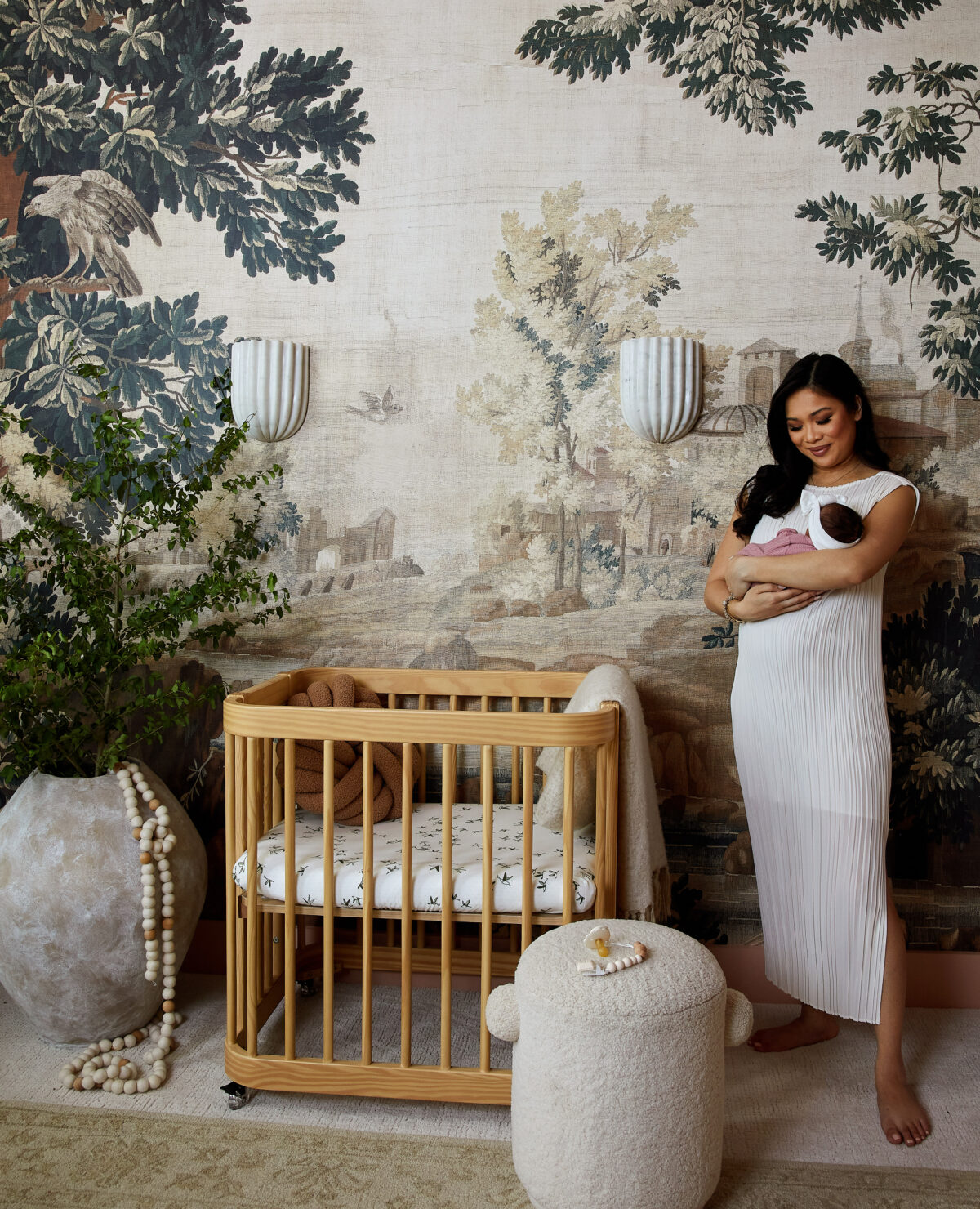 Blogger Hoang-Kim Cung holding her daughter in her nursery with mural wallpaper, nestig crib and marble sconces designed by Urbanology Designs