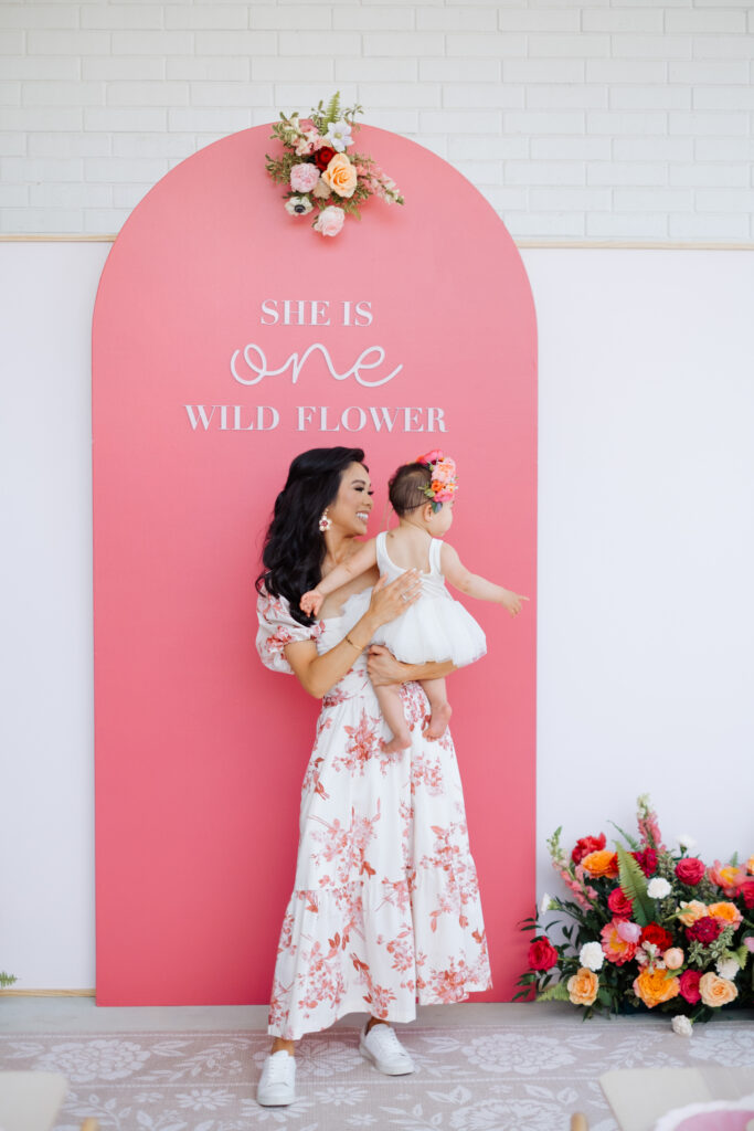 Blogger Hoang-Kim and her daughter in front of a She is one wild flower party backdrop by Glo Party Co with wood lettering and a floral cluster from Something Pretty Floral